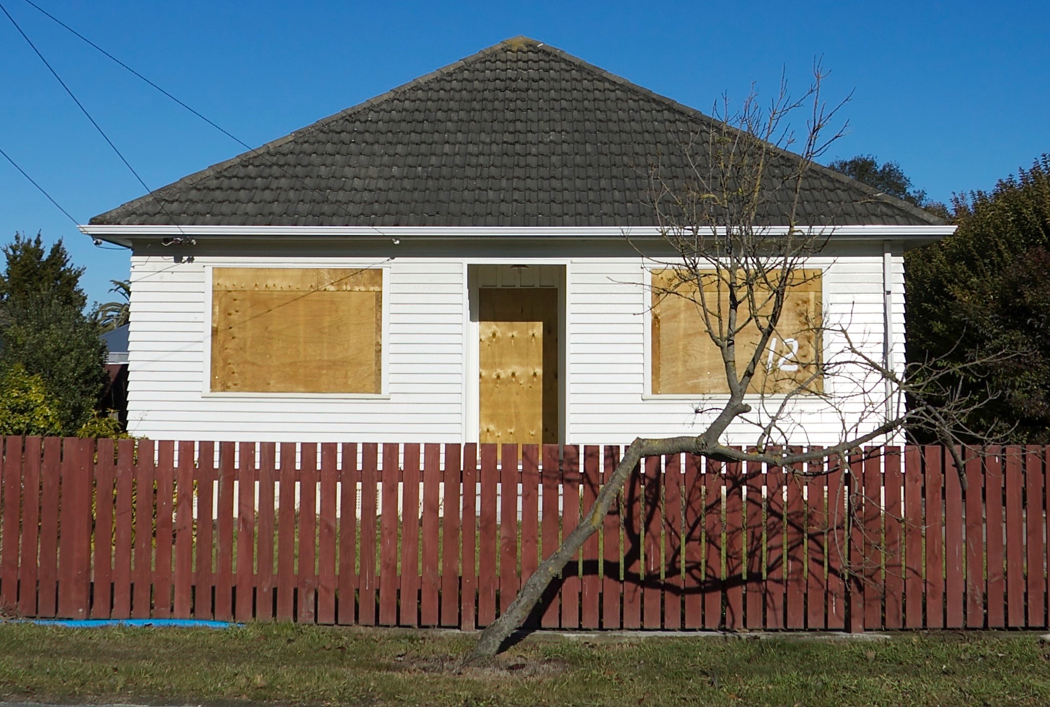 window_damage Boarded-up white house with plywood over the windows and door, red fence, and a leafless tree leaning in front under a blue sky.